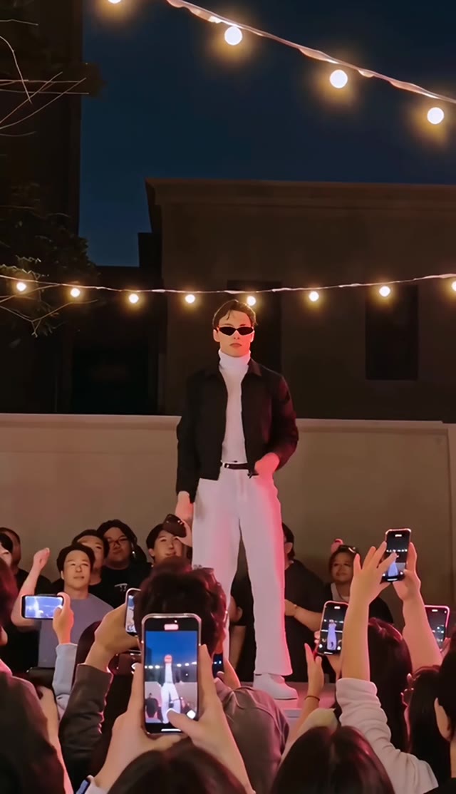 A dancer in sunglasses and a dark jacket stands beneath warm string lights.